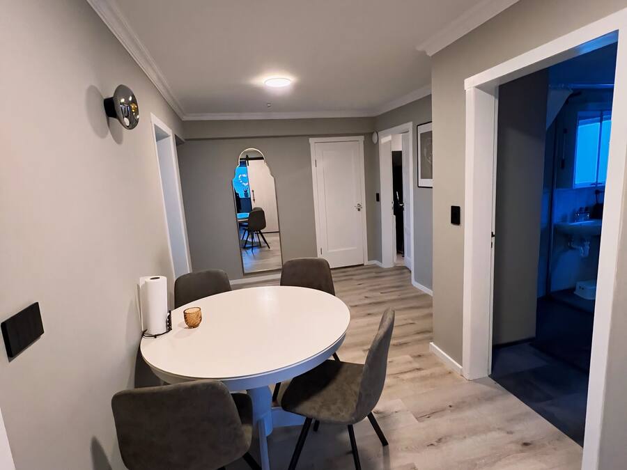 Un comedor moderno con una mesa redonda y cuatro sillas, un aplique de pared y un pasillo que conduce a otras habitaciones.