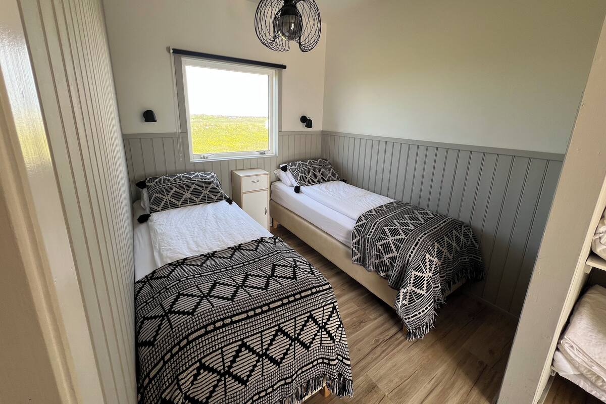 A small, compact bedroom with two single beds, a bedside table, a window, and a hanging light fixture.