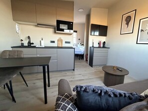 Cocina moderna con microondas, pileta y alacenas. Hay una mesa de comedor con sillas y una mesita redonda con un objeto decorativo. Un sofá con un cojín estampado está en primer plano.