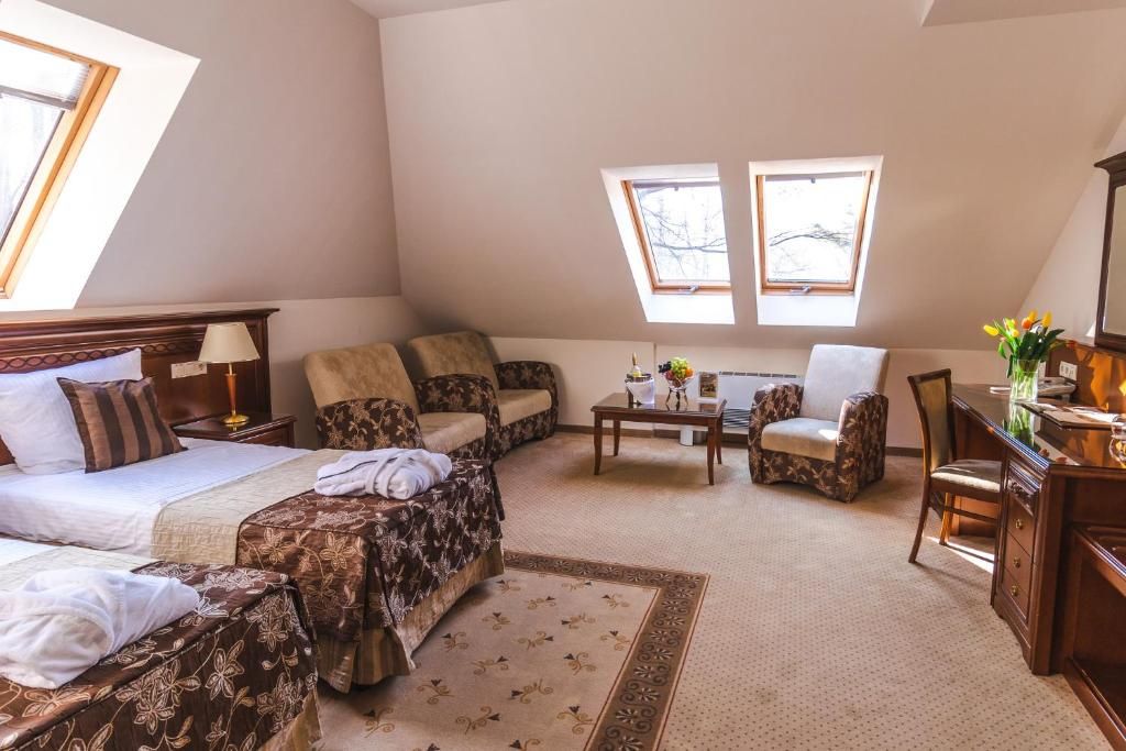 A room with a bed, two sofas, a chair, a small table, and a desk with a lamp. There are two windows providing natural light.