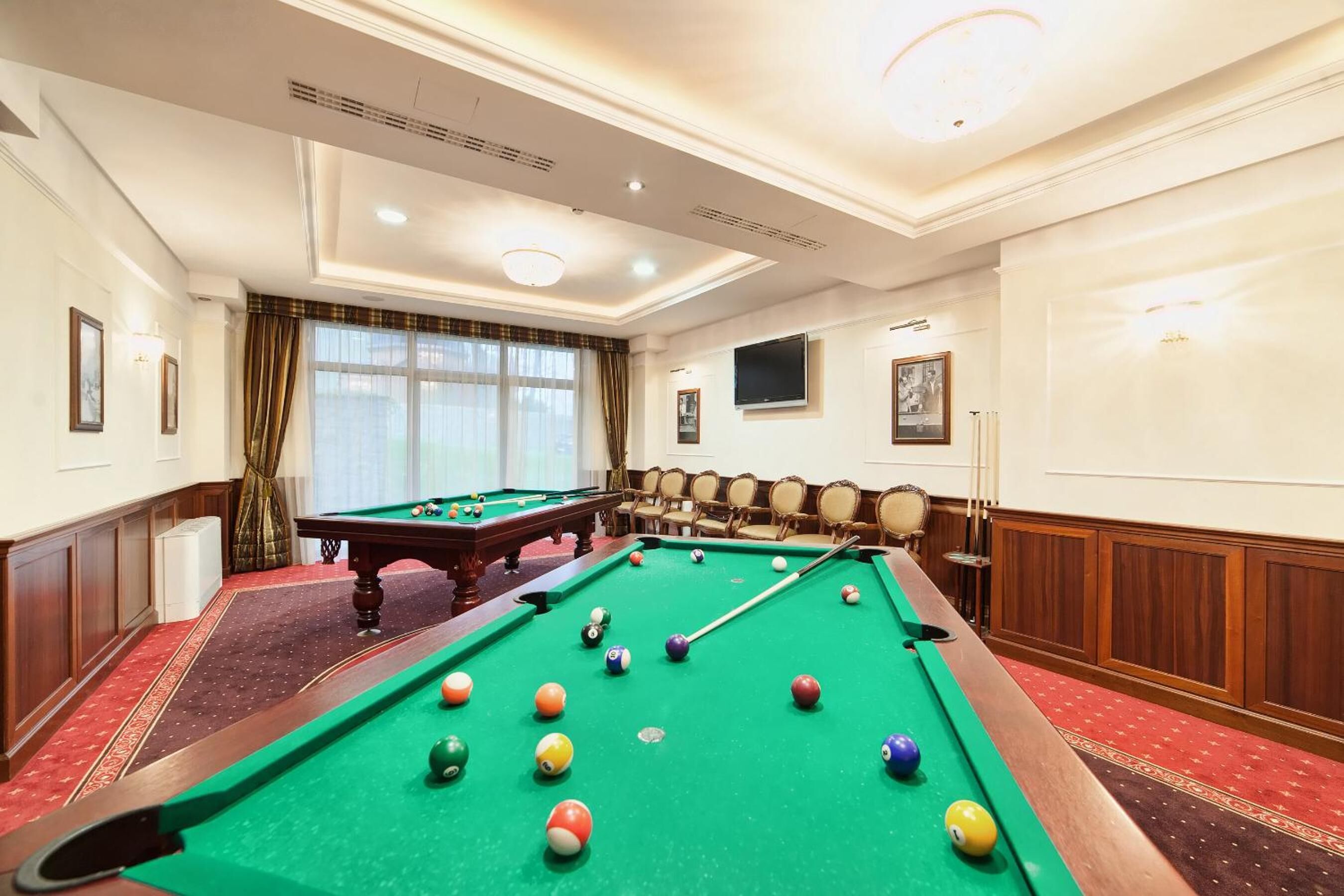 A pool table in a hotel room with a red carpet, a large window with curtains, and a television on the wall.