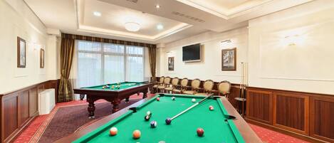 A pool table in a hotel room with a red carpet, a large window with curtains, and a television on the wall.