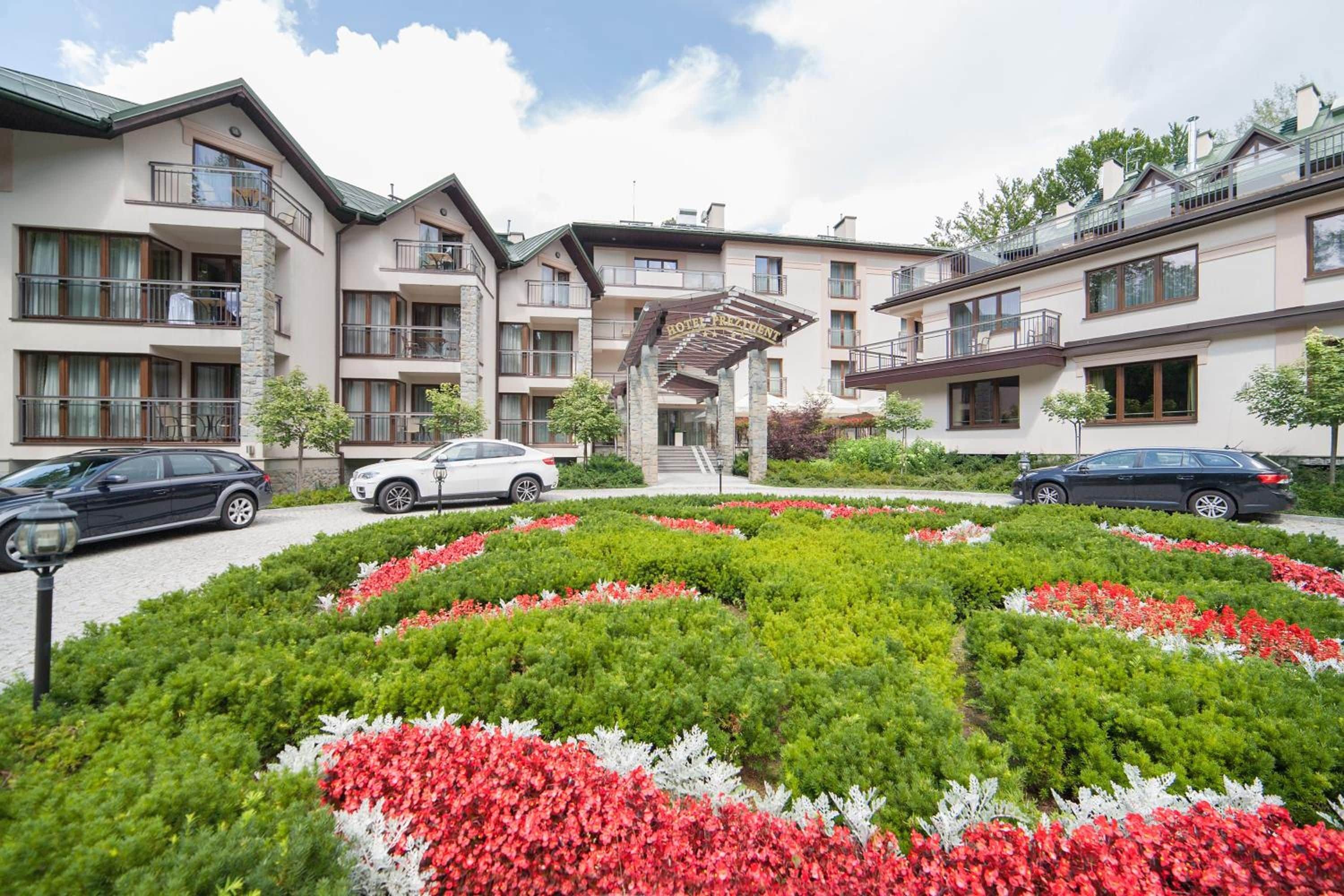 Un hotel con varios pisos, estacionamiento exterior y un jardín central con flores rojas y blancas.