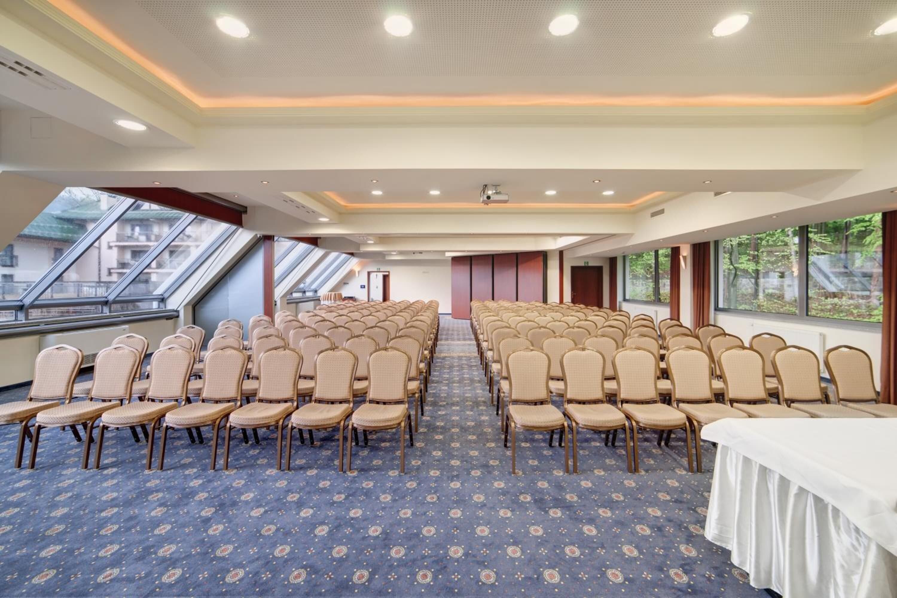 Un salón de conferencias con filas de sillas, un proyector y ventanas amplias.