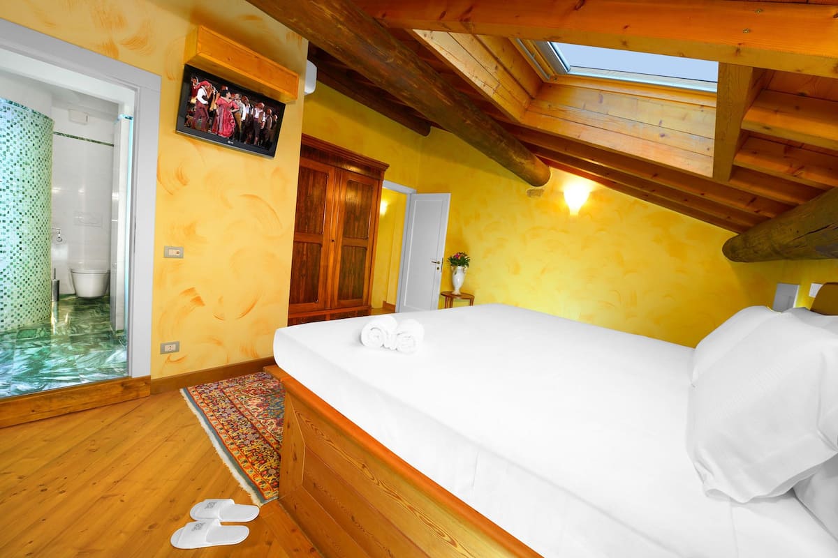 A room with a wooden ceiling, a bed with white bedding, a small table with a vase, and a colorful rug.