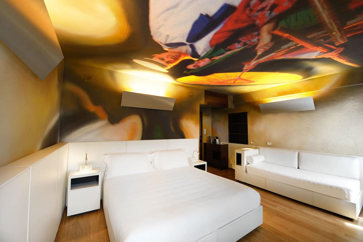 A modern hotel room with a large bed, a sofa, a nightstand, and a wall-mounted TV.
