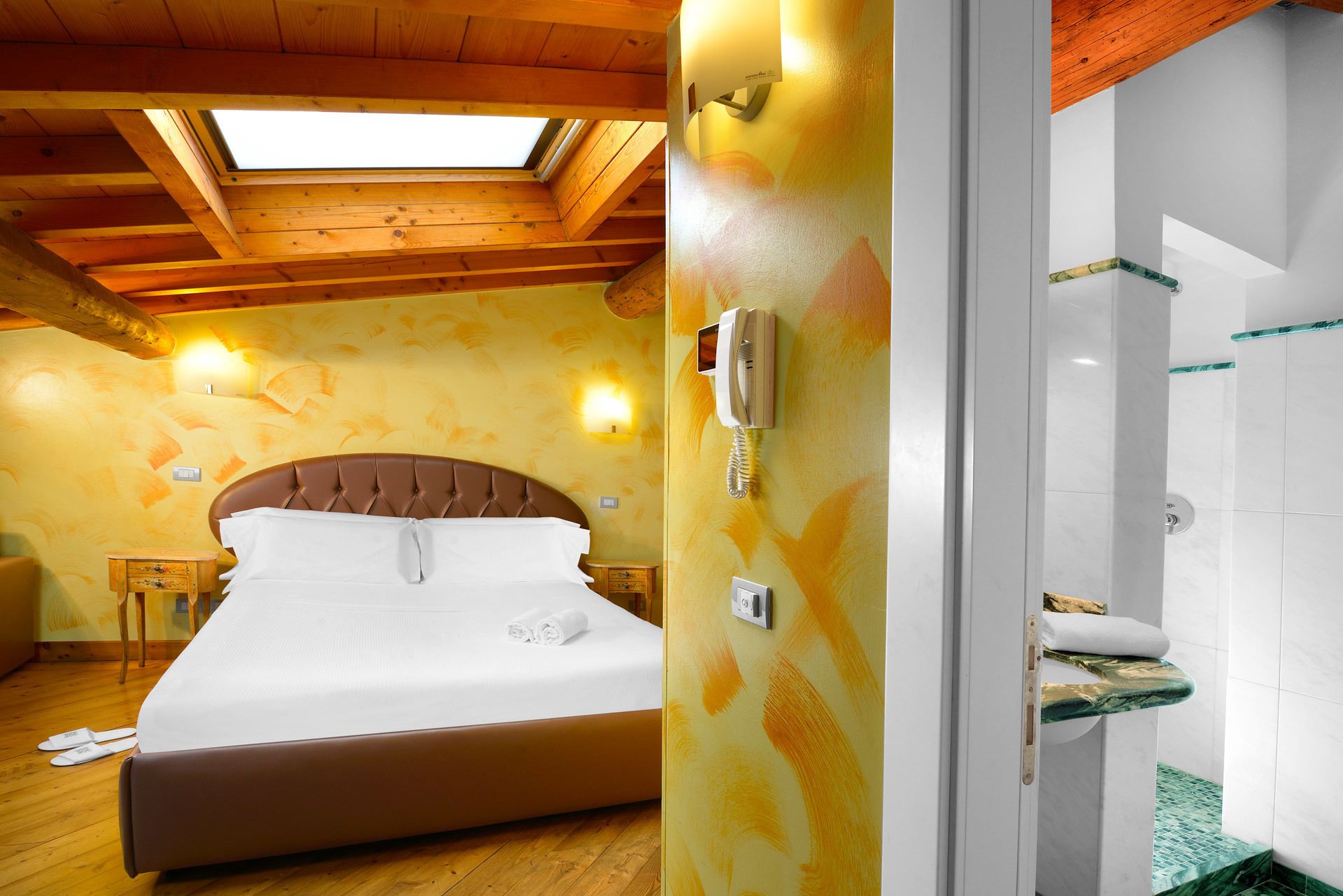 A hotel room with a bed, a wooden ceiling, a skylight, a wall-mounted phone, and a bathroom with a shower and a towel rack.