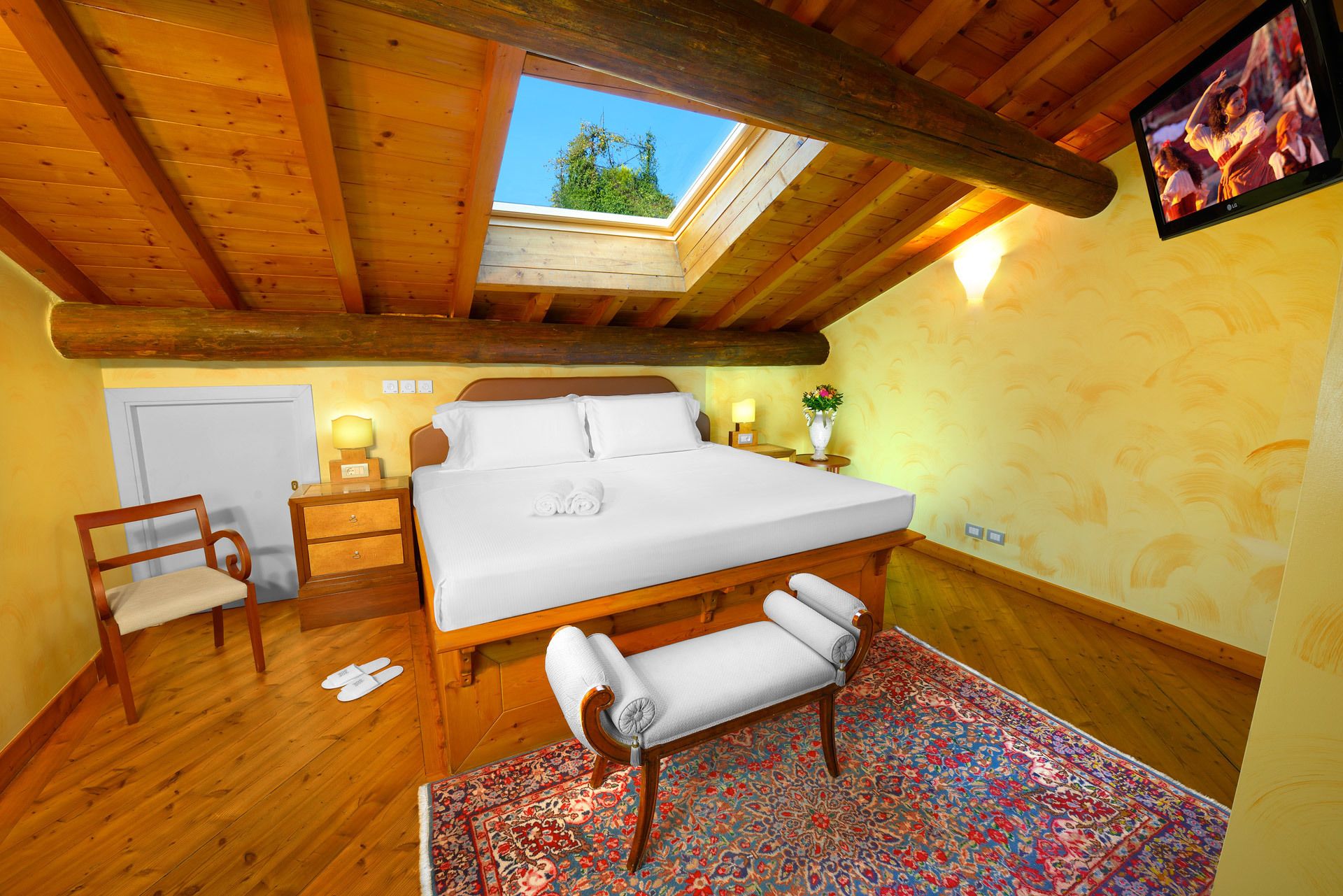 A bedroom with a large bed, a bench, a chair, a nightstand, a lamp, and a TV.
