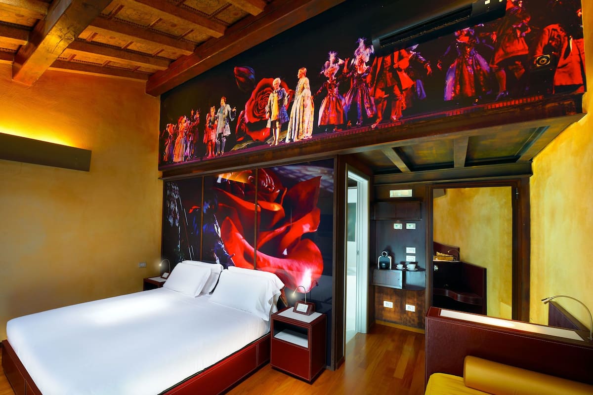 A hotel room with a large bed, a nightstand, a red bedside table, a mirror, a small seating area, and a wall with a large, colorful mural.