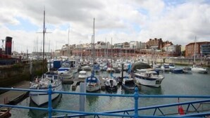 Un port de plaisance où sont amarrés de nombreux bateaux, une rambarde bleue au premier plan et des bâtiments en arrière-plan.