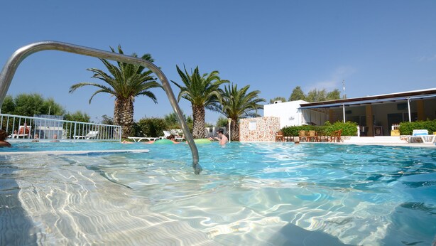 A clear swimming pool with a metal arch entrance, palm trees, and a building with outdoor seating. - Aslanis Village Hotel (Kos)