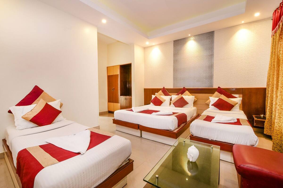 Habitación de hotel con cuatro camas dispuestas en forma de U, una mesa de centro de vidrio, una silla de cuero rojo y un cuadro abstracto grande en la pared.