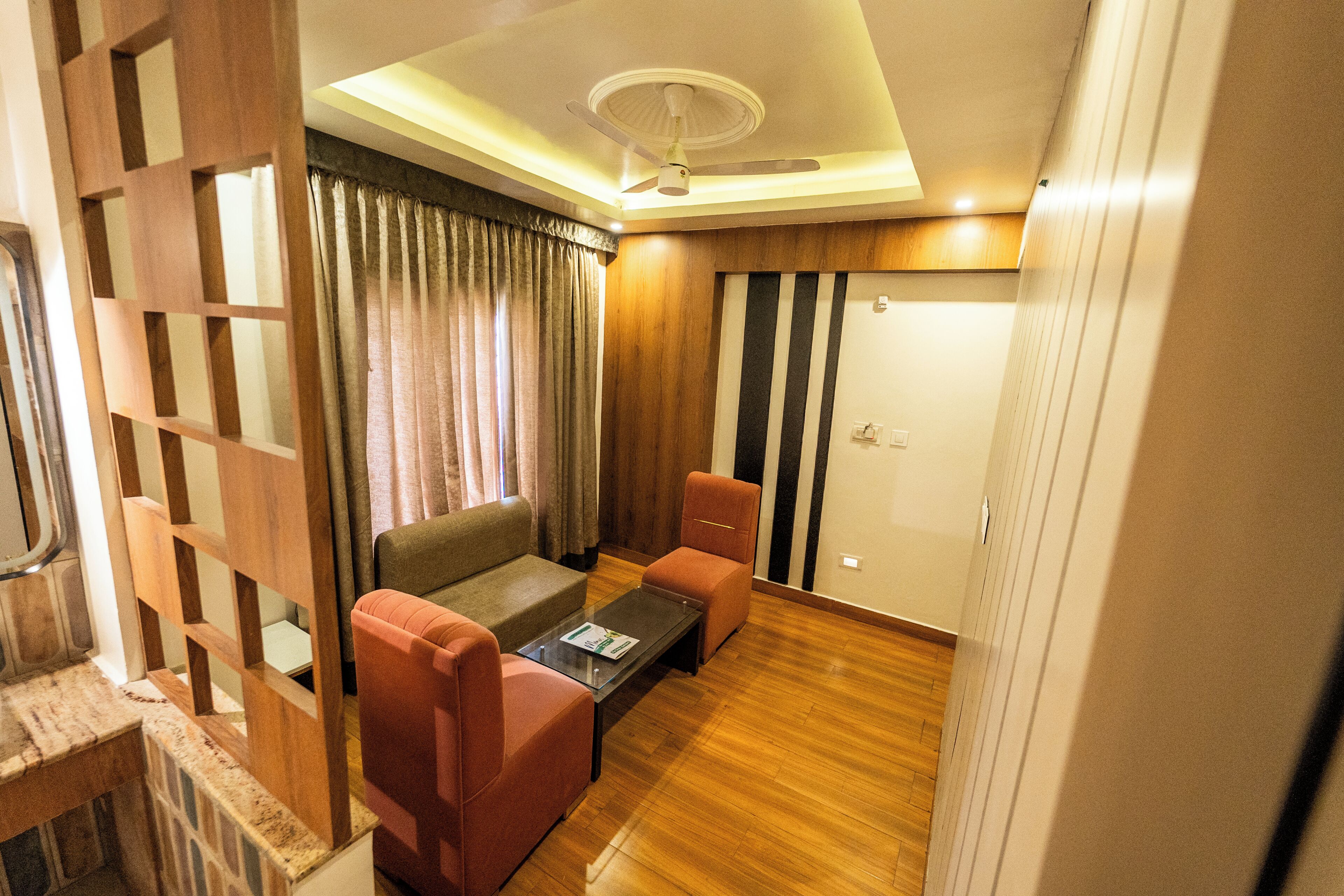 Una habitación con sofá, dos sillones, una mesa de centro, ventilador de techo, suelo de madera y una puerta con franjas verticales.