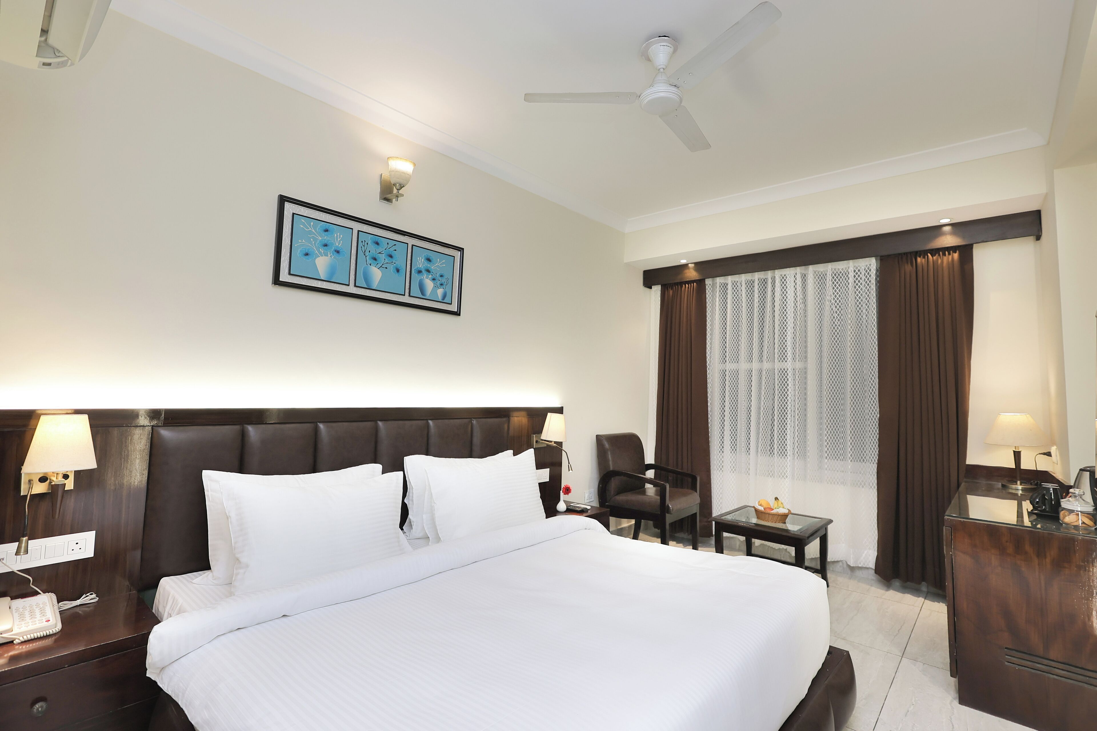 Une chambre d’hôtel avec un grand lit, un bureau, une chaise, une petite table, un ventilateur de plafond et une fenêtre avec des rideaux.
