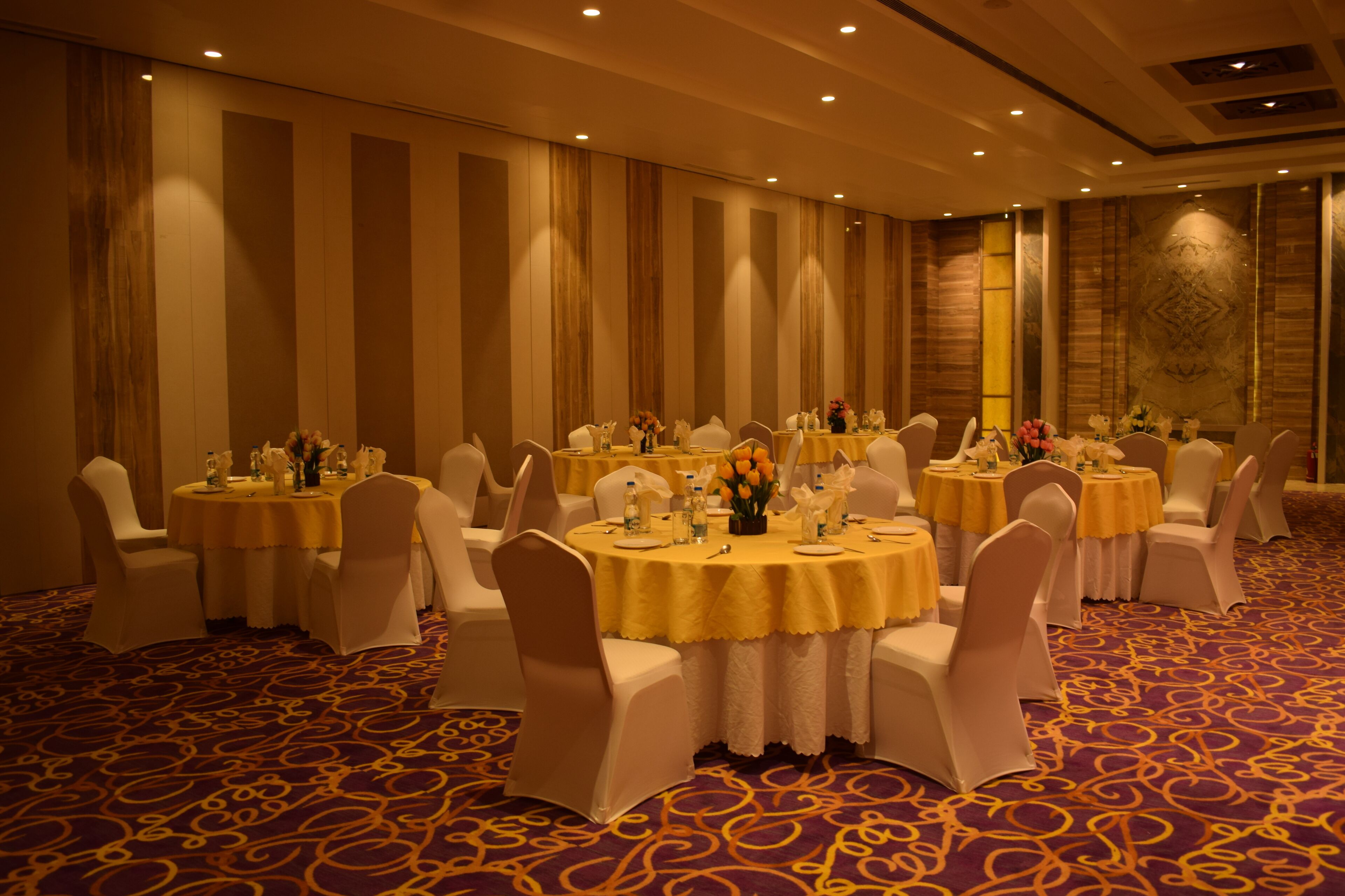 Un salón de banquetes con mesas redondas, sillas blancas y manteles amarillos.