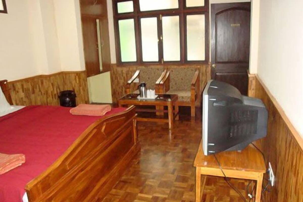 A room with a bed, a wooden table with a television, a chair, and a window with blinds.