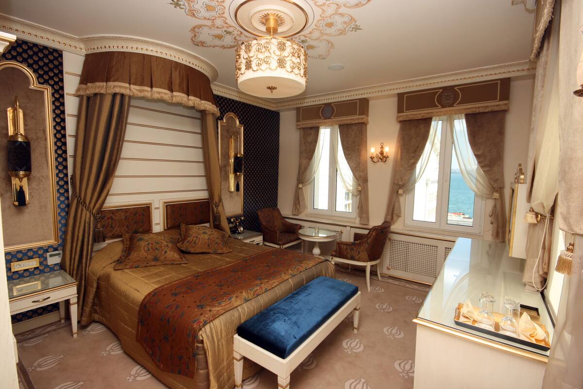 A luxurious bedroom with a large bed, a bench, a small table, and a view of the sea through the windows.