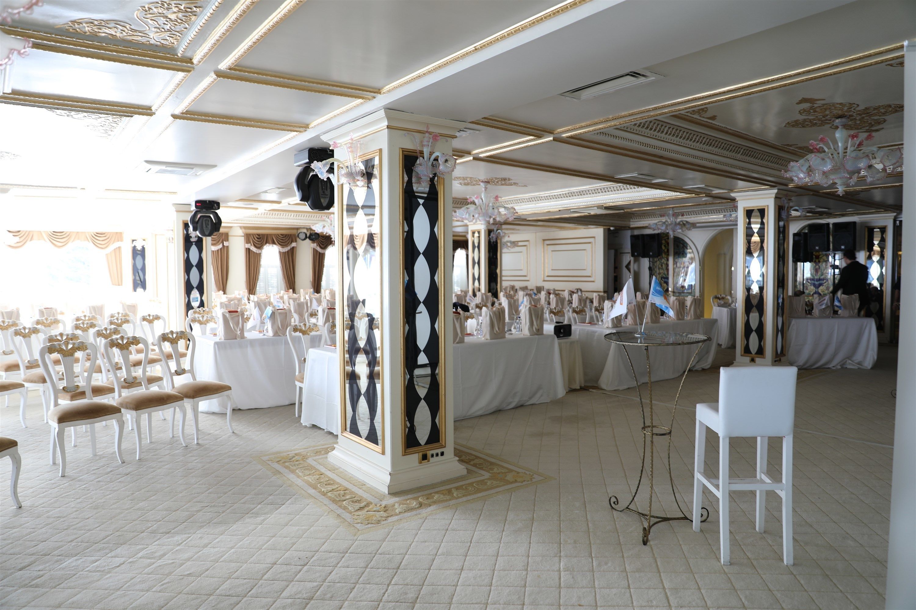 A banquet hall with white tables and chairs, ornate columns, and a chandelier.