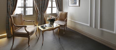 A room with two ornate chairs, a small table with a vase, and a framed picture on the wall.