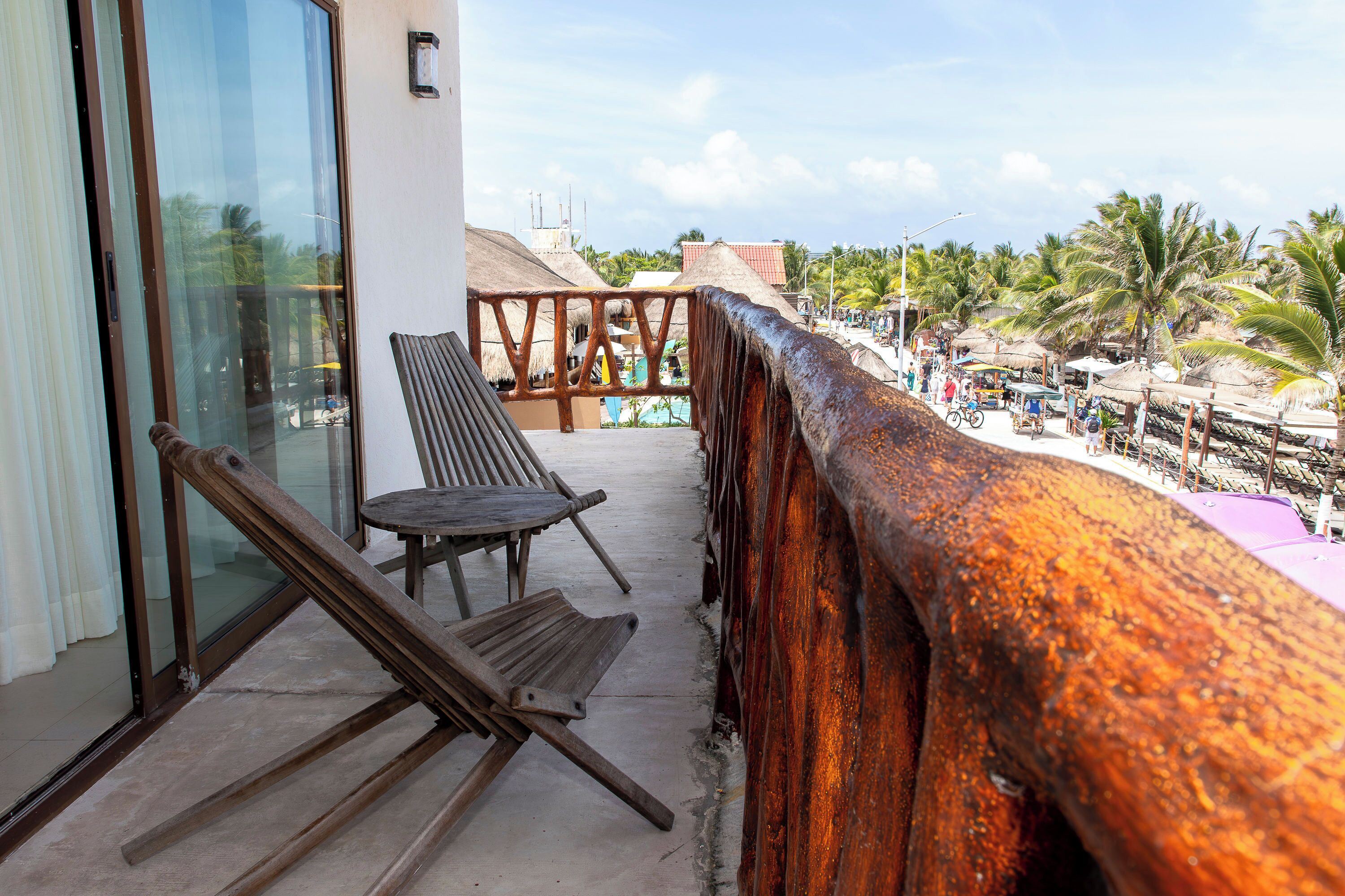 A balcony with a wooden bench, a glass door, and a view of a beach with palm trees and a thatched-roof building.
