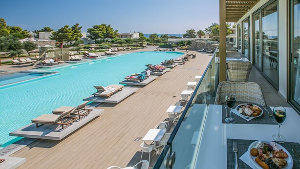 A rooftop pool area with lounge chairs and tables, overlooking a swimming pool and a scenic view. - Almyra Resort (Ierapetra)
