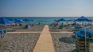 A beach with a wooden walkway leading to the sea, surrounded by blue umbrellas and beach chairs. - Almyra Resort (Ierapetra)