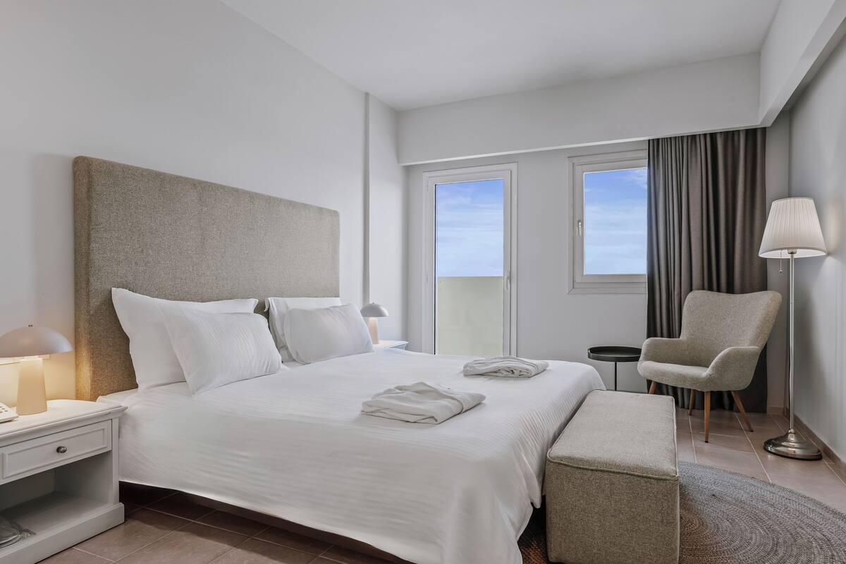 Ein modernes Hotelzimmer mit einem großen Bett, einem Nachttisch mit Lampe, einem Sessel und Blick auf den Himmel durch ein Fenster.