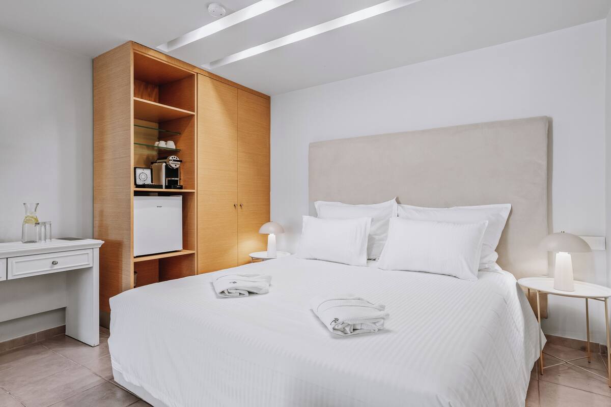 Ein modernes Hotelzimmer mit einem großen Bett, einem Holzschrank, einem Schreibtisch mit Lampe und einem kleinen Tisch ebenfalls mit Lampe.
