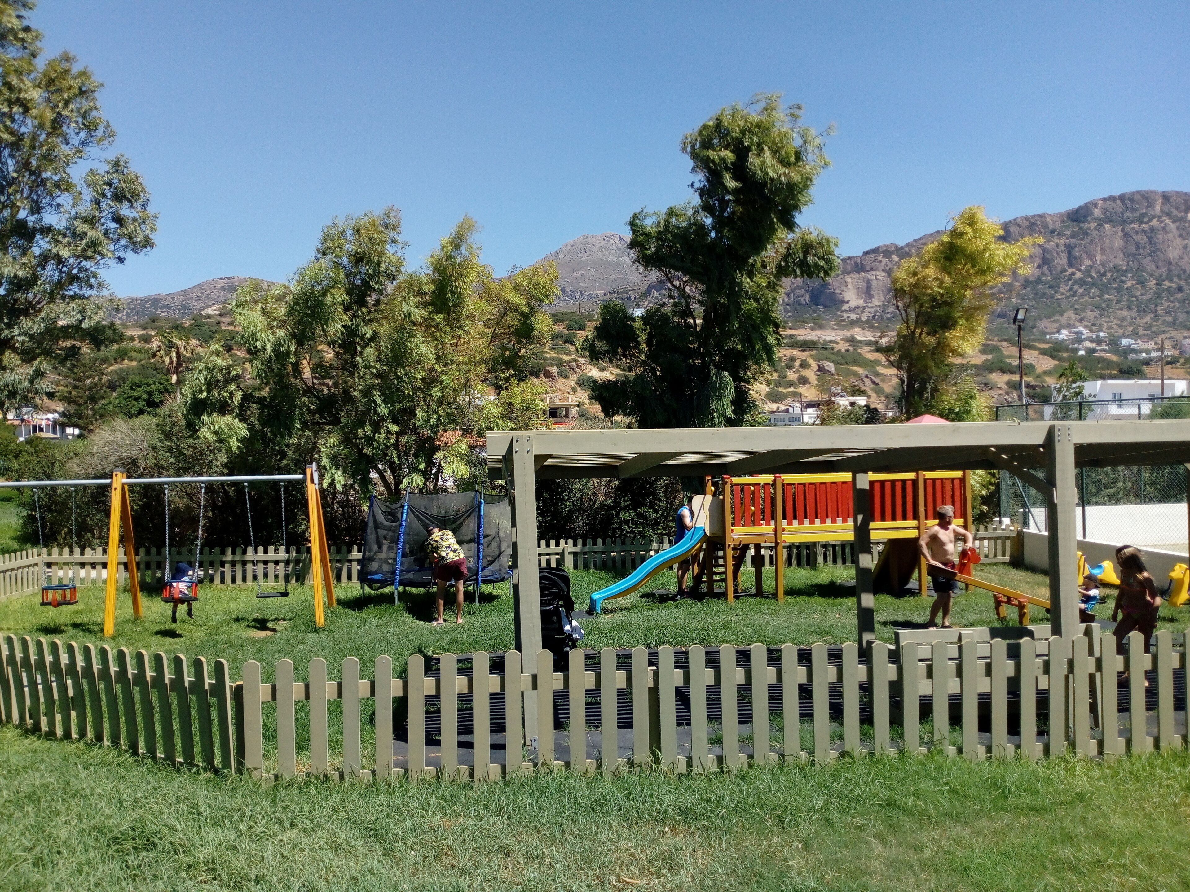 Un parque infantil con tobogán, columpios y estructuras para escalar, rodeado por una valla de madera y árboles.