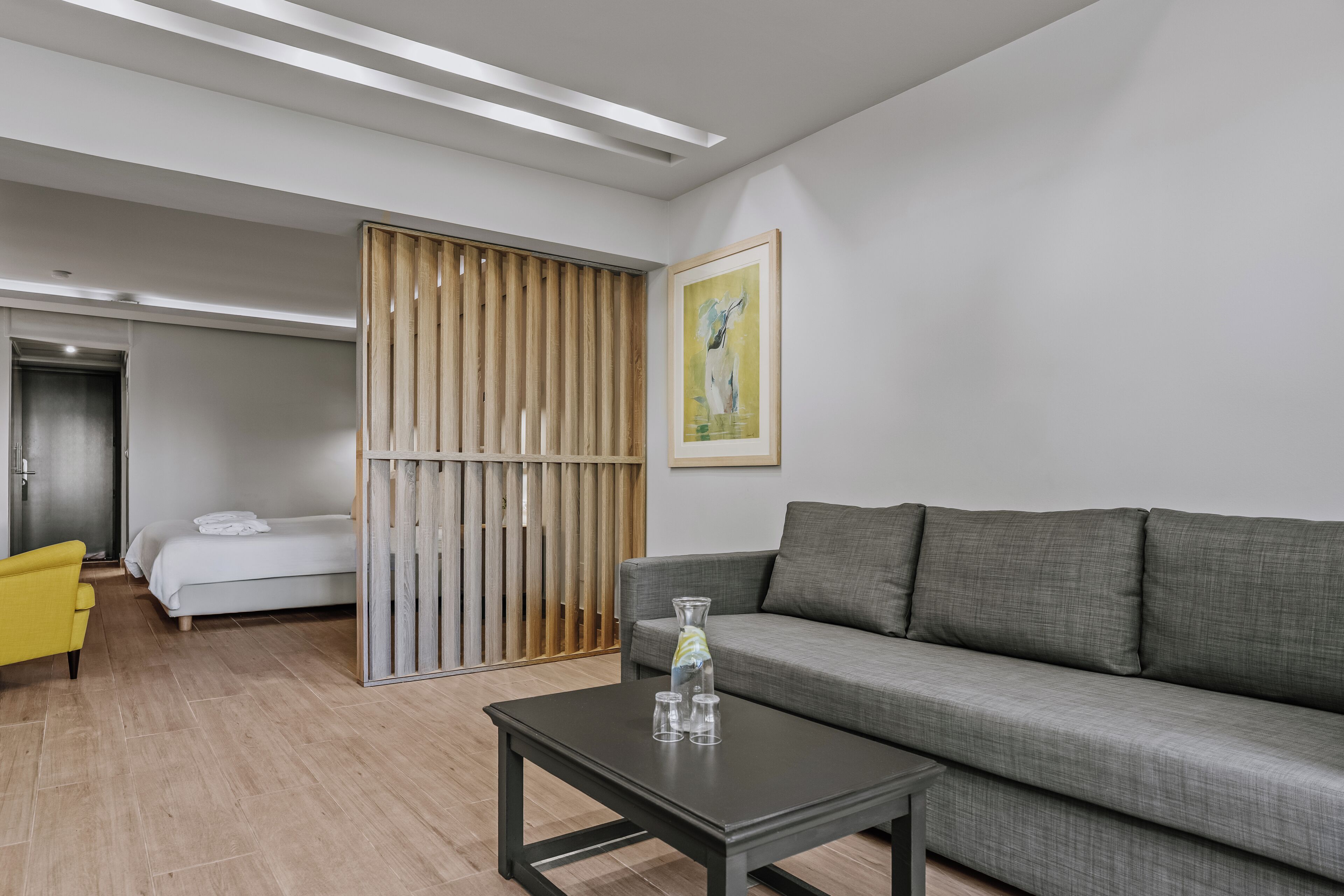 A modern hotel room with a grey sofa, a small table with glasses, a yellow armchair, and a bed with white linens.