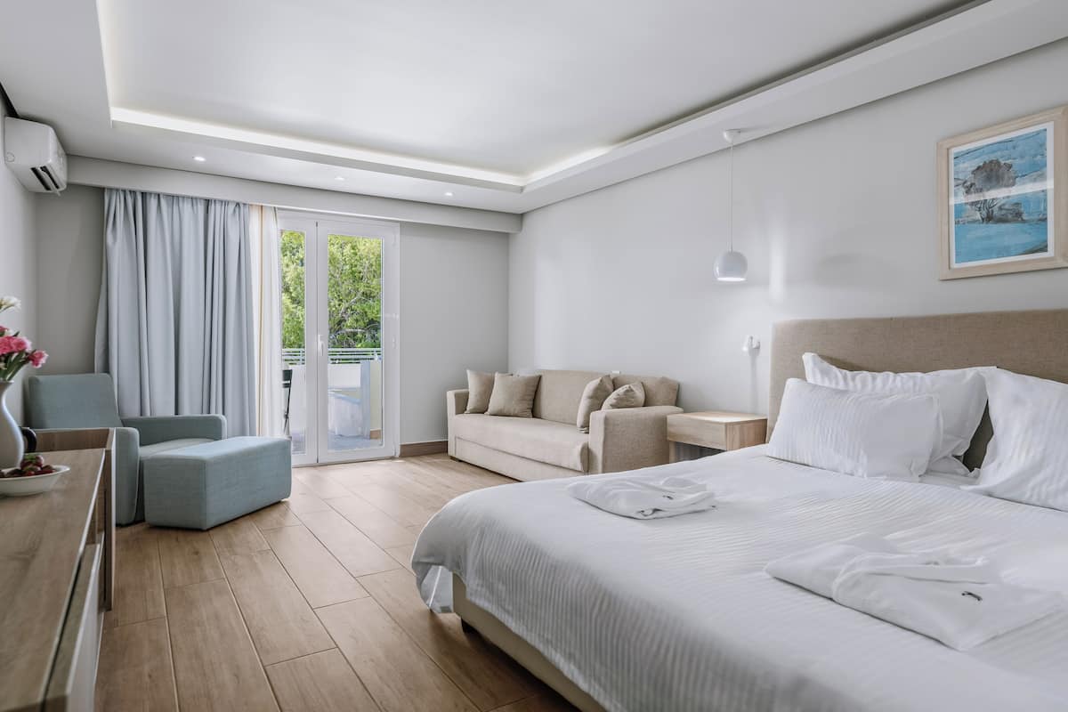 Ein modernes Hotelzimmer mit einem großen Bett, einer Couch, einem Schreibtisch und Blick auf Grünanlagen durch die Schiebeglastür.