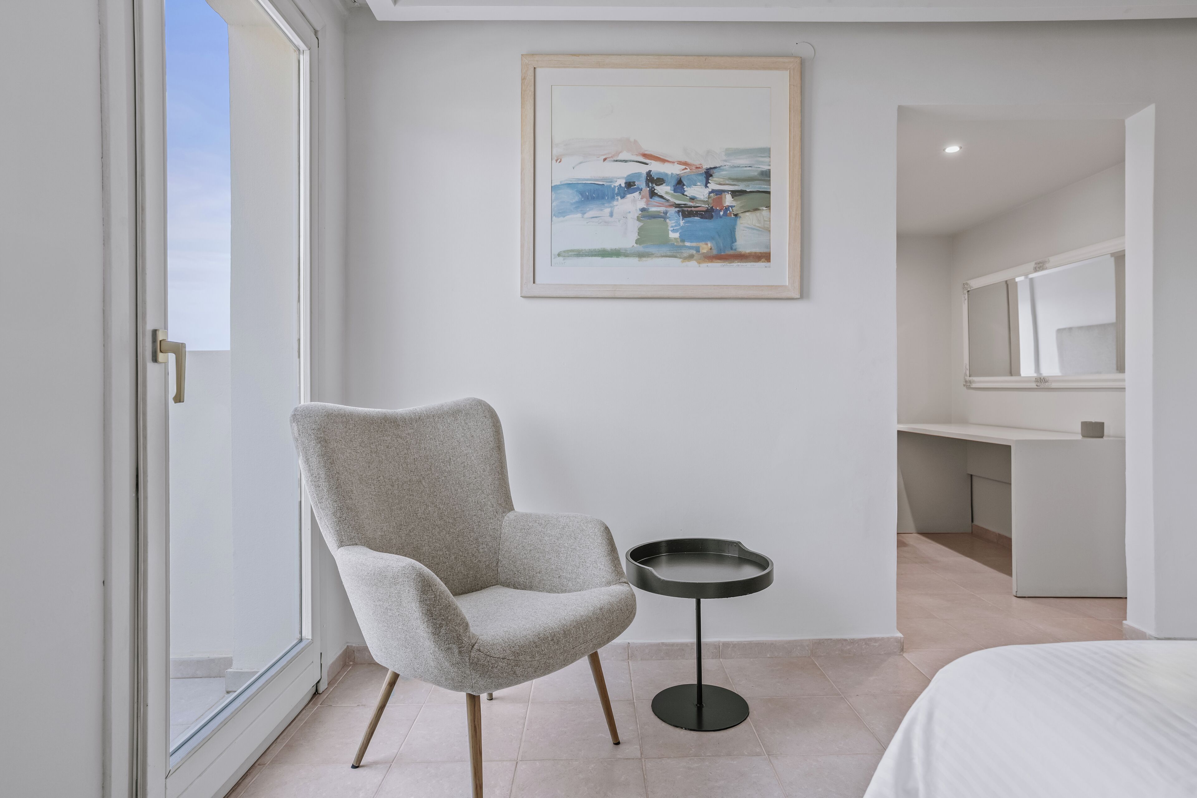 A modern hotel room with a large window, a grey armchair, a small round table, and a painting on the wall.