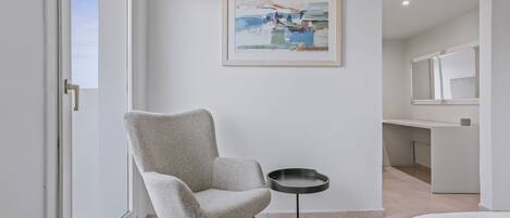 A modern hotel room with a large window, a grey armchair, a small round table, and a painting on the wall.