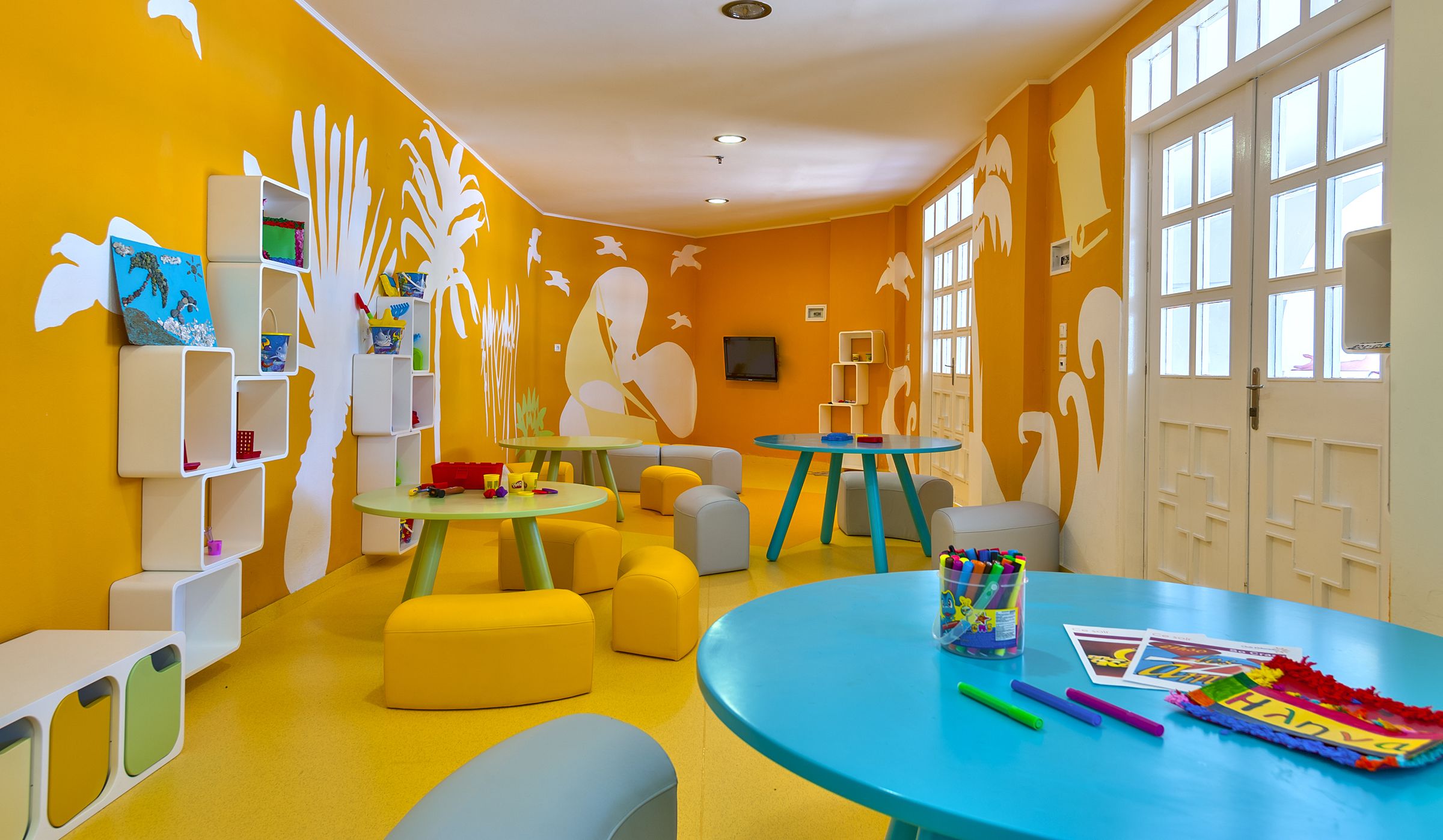 A brightly colored children's playroom with yellow walls, white shelves, and various play furniture.