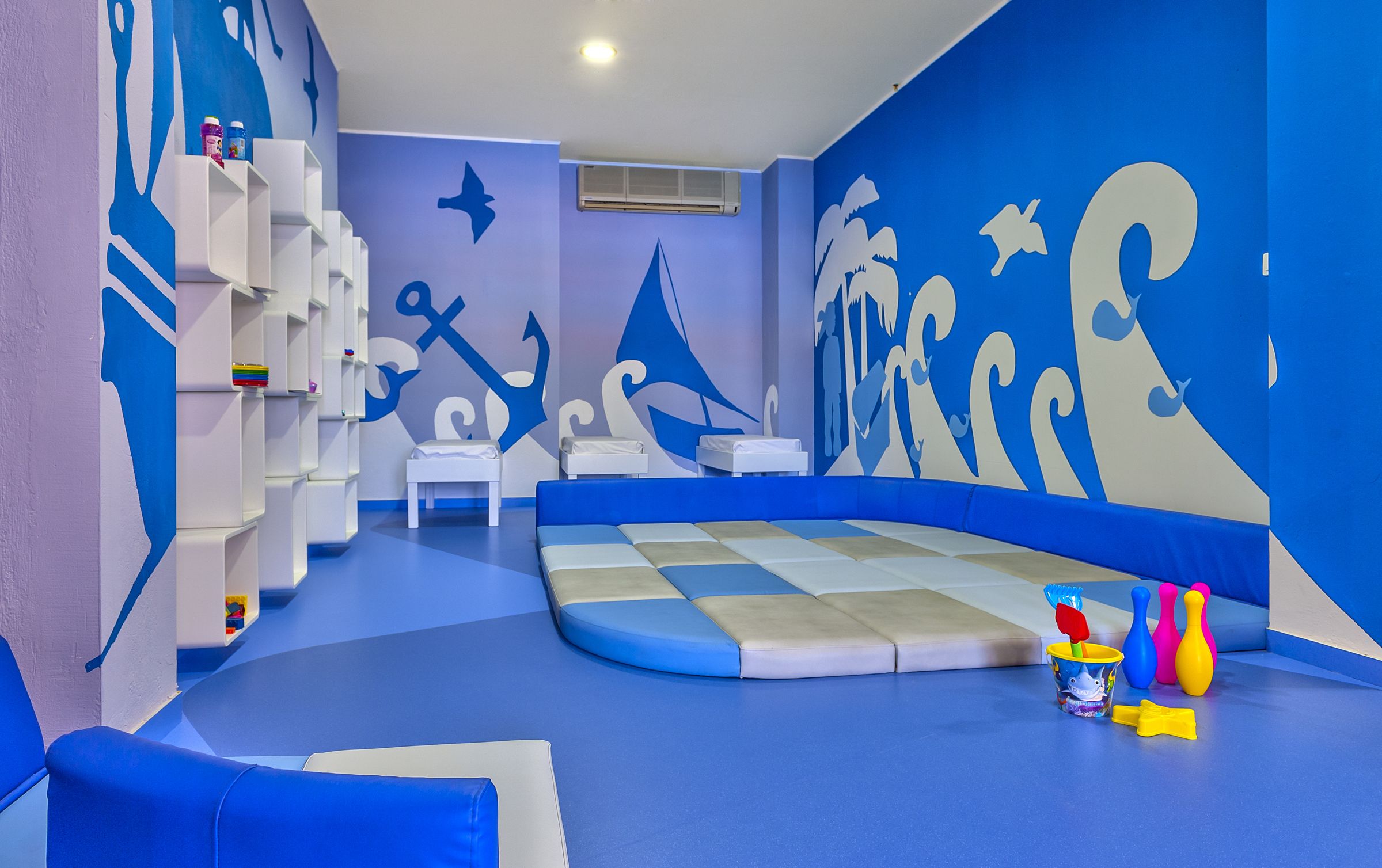Zona de juegos infantiles con una decoración azul y blanca, que incluye suelo acolchado, zonas de estar y una pared con motivos náuticos.