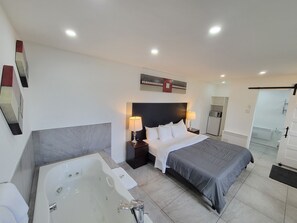 Quarto de hotel moderno com uma cama grande, banheiro separado com banheira, uma pequena escrivaninha e uma geladeira.