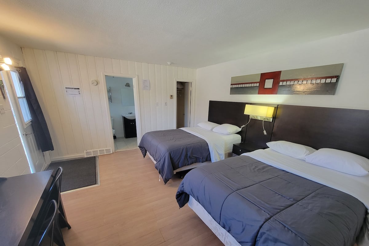 A hotel room with two beds, a desk, a chair, a bathroom visible through an open door, and a wall-mounted light fixture.