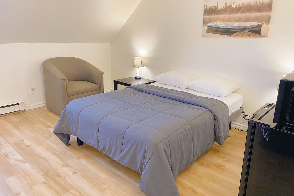 A bedroom with a bed, a chair, a desk with a lamp, and a TV.