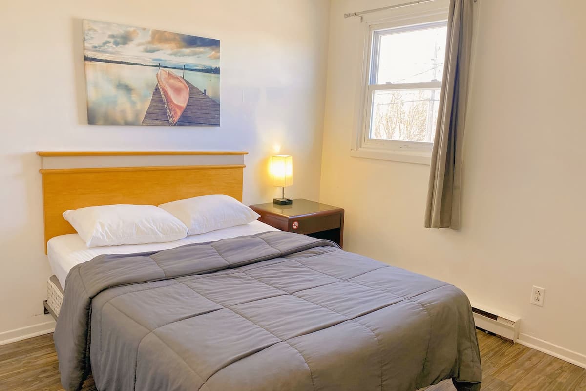 A neatly made bed with a grey duvet, a wooden headboard, a nightstand with a lamp, a painting of a boat on a pier, and a window with curtains.