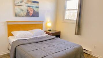 Cama bem arrumada com edredom cinza, cabeceira de madeira, mesa de cabeceira com abajur, quadro de um barco em um cais e janela com cortinas.
