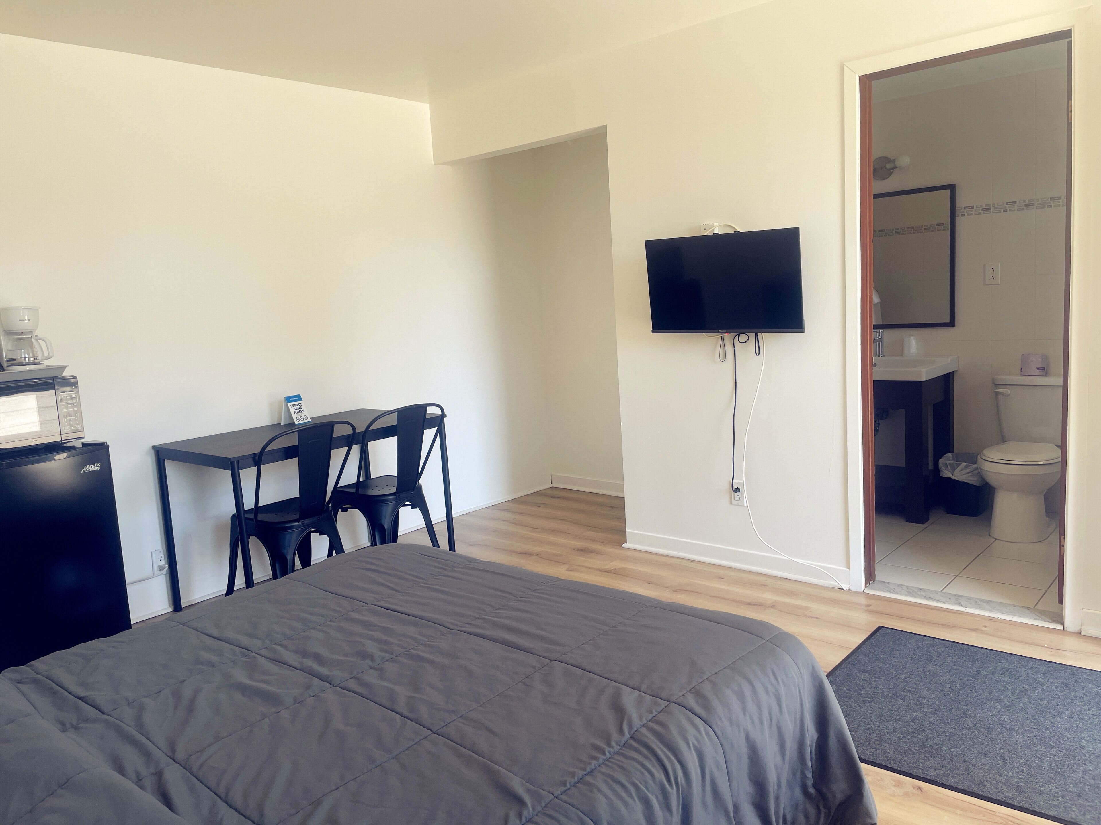 Una habitación de hotel con una cama, un escritorio con dos sillas, un televisor montado en la pared y un baño visible a través de una puerta abierta.