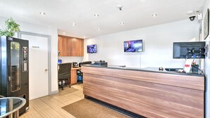 A modern hotel reception area with a wooden reception desk, a glass partition, a seating area with a chair, and a security camera on the ceiling. - Motel Grande ile (Salaberry De Valleyfield)
