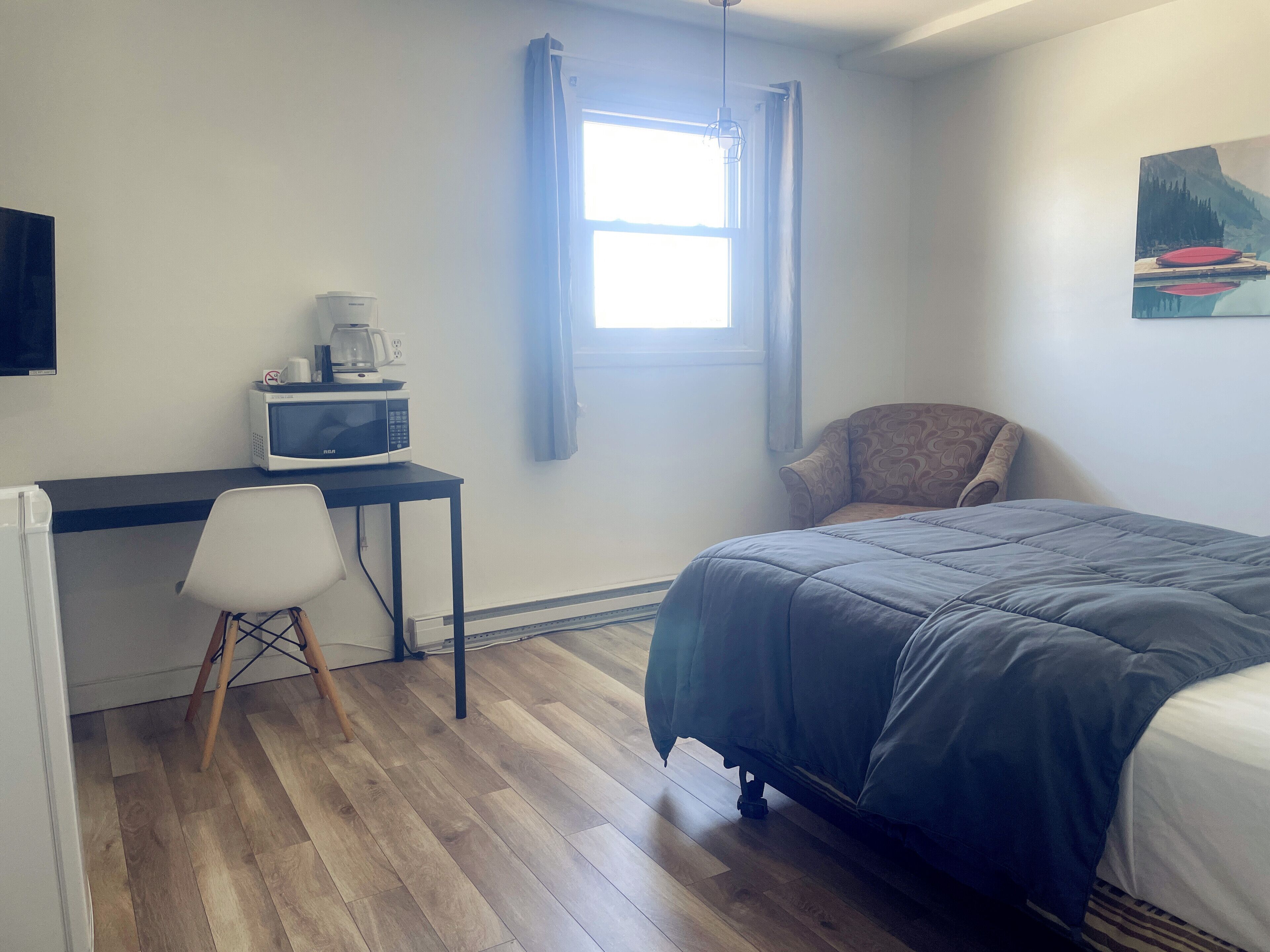 Une chambre à coucher avec un lit, un bureau avec un ordinateur et une machine à café, une chaise et une fenêtre avec des rideaux.