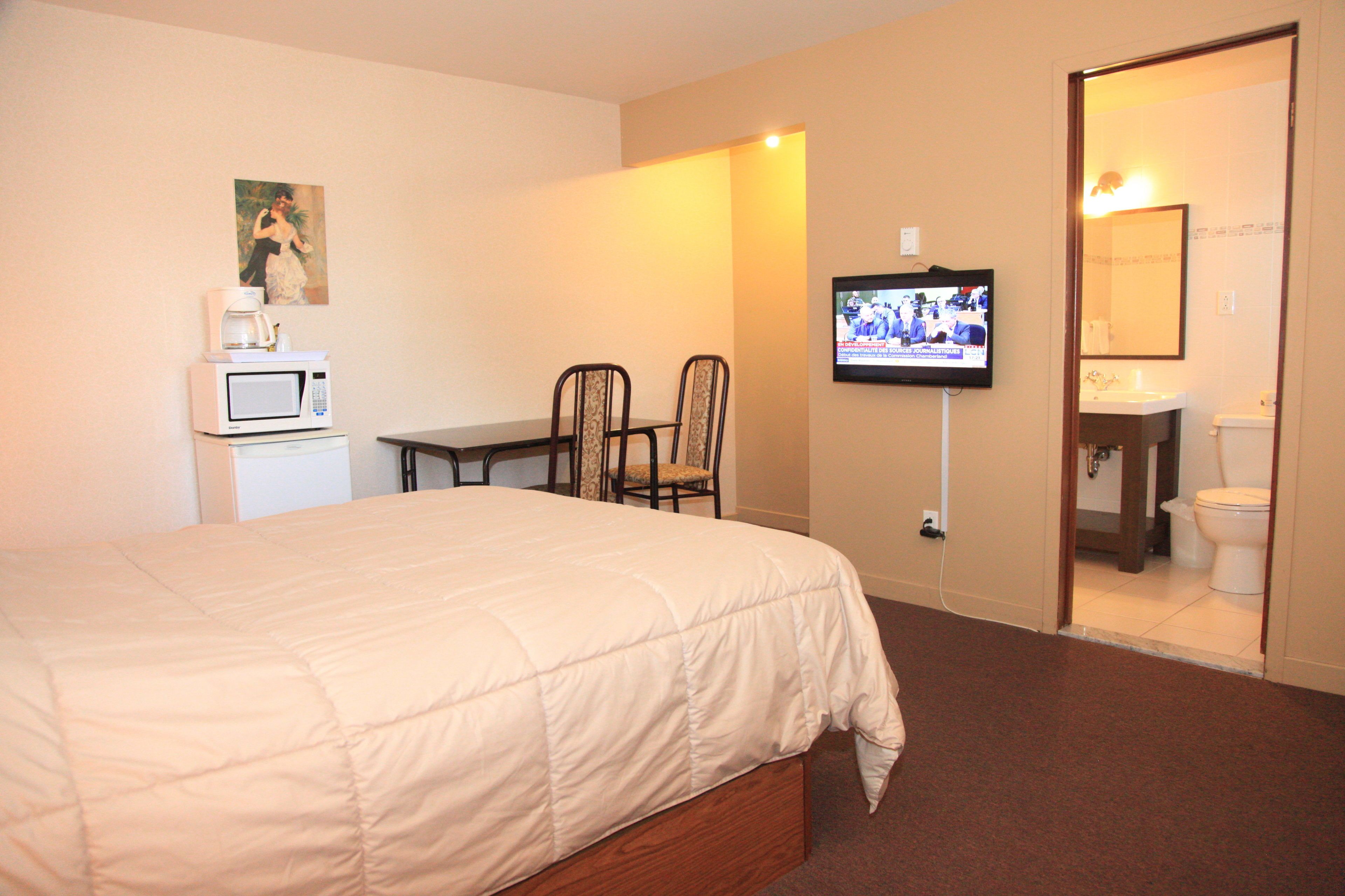 Habitación de hotel con cama, televisor montado en la pared, una pequeña mesa con sillas, microondas y baño visible a través de una puerta abierta.