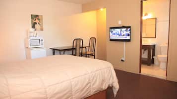 Habitación de hotel con cama, televisor montado en la pared, una pequeña mesa con sillas, microondas y baño visible a través de una puerta abierta.