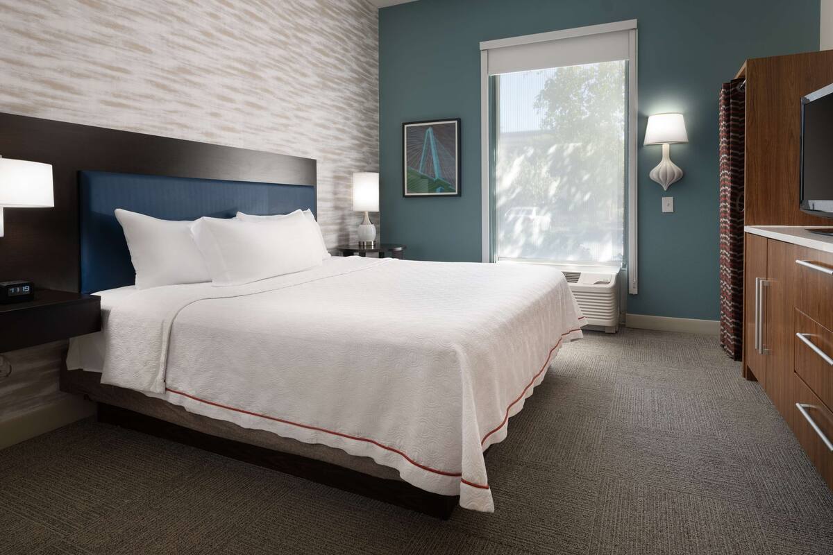 A hotel room with a large bed, a headboard, a nightstand with a lamp, a window with a view, and a TV on a wooden cabinet.