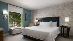 Une chambre d’hôtel avec un grand lit, deux tables de chevet avec des lampes, une fenêtre avec un climatiseur et un tableau accroché au mur.