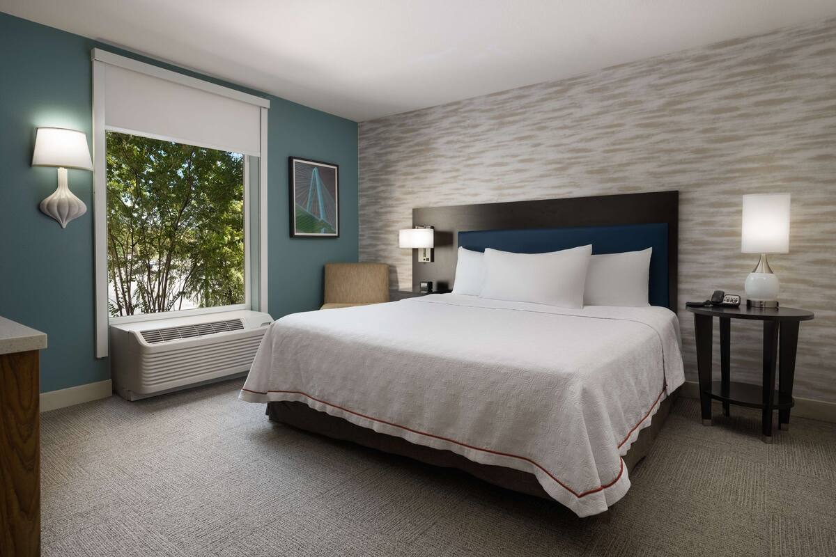 A hotel room with a large bed, two bedside tables with lamps, a window with an air conditioner, and a wall-mounted picture.