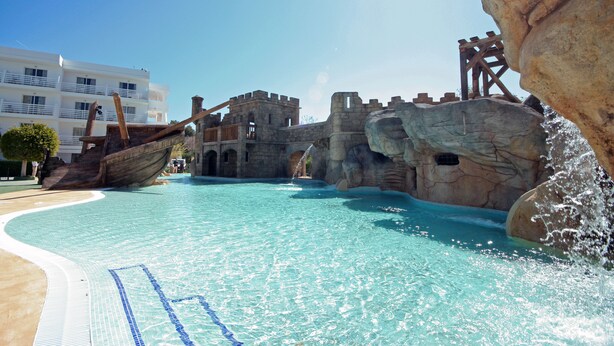 A swimming pool with a shipwreck theme, clear blue water, and a castle-like structure in the background. - Pirates Village (Calvia)
