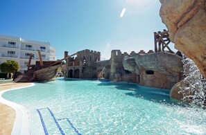 A swimming pool with a shipwreck theme, clear blue water, and a castle-like structure in the background. - Pirates Village (Calvia)
